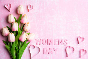 Womens day concept. Pink tulips and paper hearts with Wooden letters forming word Womens day written on pink and white wooden background.