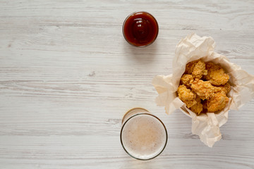 Chicken bites in paper box, barbecue sauce and glass of beer on a white wooden table, top view. Flat lay, overhead, from above. Copy space.