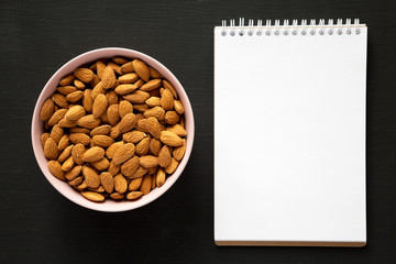 Full pink bowl of almonds, blank notepad on black background, overhead view. Flat lay, from above, top view. Space for text.