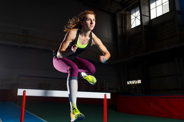 Young sportswoman running and jumping over hurdle