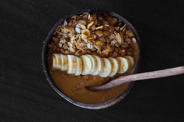 Healthy vegan breakfast with cereals, nuts and banana in original handmade coconut bowl with wooden spoon on dark background