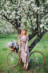 smiling blonde girl in a dress leaned against a tree near a bicycle with flowers