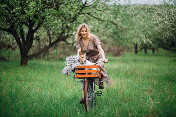 close-up blonde girl in a dress riding a bicycle with a basket with a dog in a blooming garden