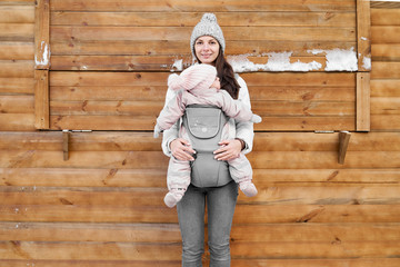 Little baby girl and her mother babywearing the ergo carrier in winter wooden background