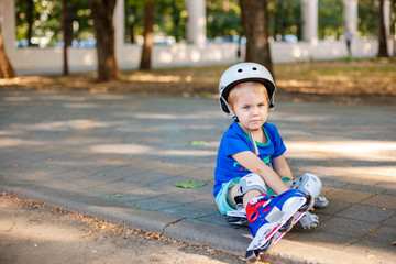 Little blonde boy 3 years old in white sport helmet and blue t-shirt riding on the roller-slates in the summer green park. Special problems with kid's eyes. Myopie, astigmatism, cross-eyed