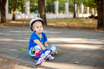 Obraz premium Mały blond chłopiec 3 lata w białym sportowym kasku i niebieskiej koszulce na rolkach w letnim zielonym parku. Specjalne problemy z oczami dziecka. Krótkowzroczność, astygmatyzm, zezowate oczy