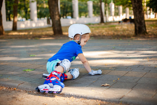 Little blonde boy 3 years old in white sport helmet and blue t-shirt riding on the roller-slates in the summer green park. Special problems with kid's eyes. Myopie, astigmatism, cross-eyed - Powered by Adobe