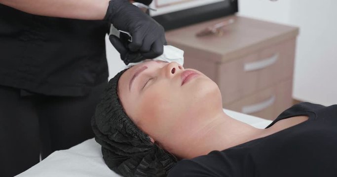 Woman cosmetologist using a clean cloth to remove paint residues from the eyebrows. A female pacient with her eyes closed in a beauty salon