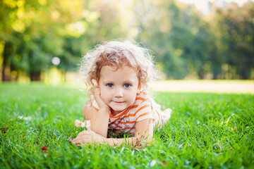 Portrait of adorable little curle girl lying on grass and propping up her face at summer green park