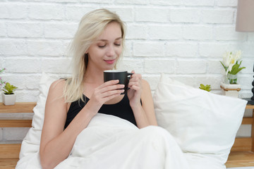 Beautiful young smiling woman sitting on bed with a cup of coffee in the morning at bedroom.