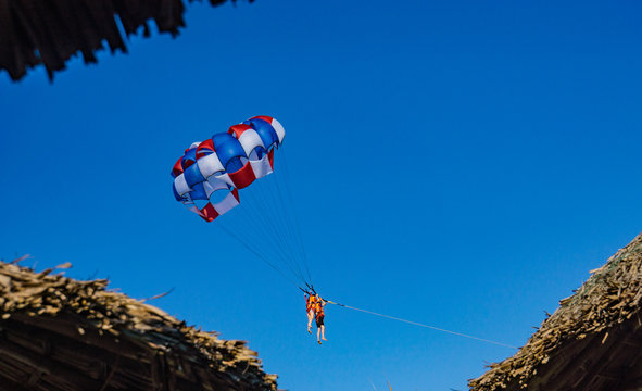 parasail with two swimmers hanging framed by the thatched roofs of the beach huts
