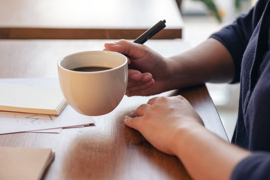 Closeup image of a woman's hand holding a cup of hot coffee while working on business document on wooden table