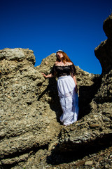 Portrait of young beautiful redhead woman in dress sitting on the rock and blue sky