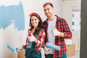 cheerful man and woman showing thumbs up while holding rollers in hands