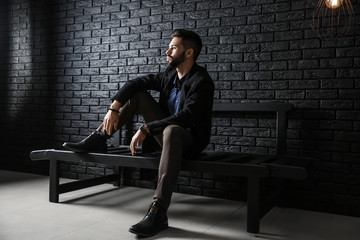 Fashionable young man sitting on bench against dark brick wall