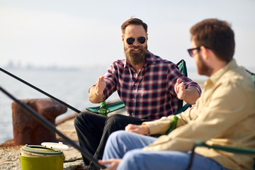 leisure and people concept - happy male friends with rods on pier at sea telling stories about...