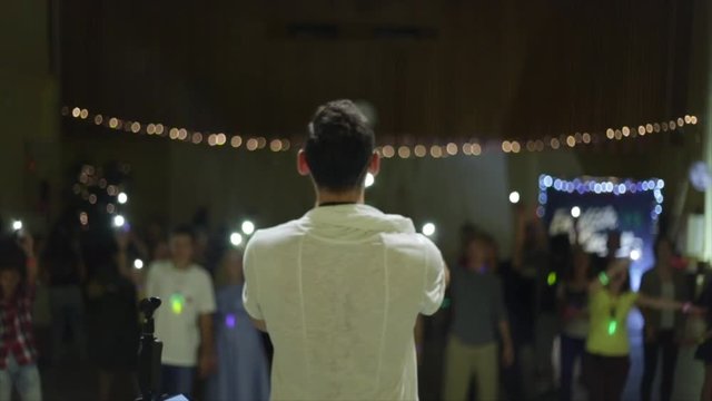 Back view of young caucasian singer against the auditorium. Back view man in white t shirt performing on stage.