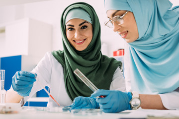 smiling female muslim scientists holding pipette and glass test tube during experiment in chemical...