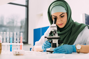 beautiful female muslim scientist looking through microscope during experiment in chemical...