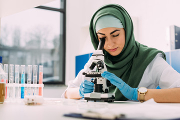 beautiful female muslim scientist looking through microscope during experiment in chemical laboratory - Powered by Adobe