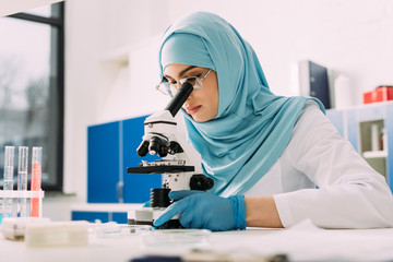 concentrated female muslim scientist looking through microscope during experiment in chemical laboratory