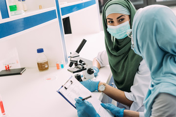 female muslim scientists in hijab using microscope and clipboard during experiment in chemical...