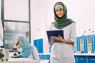female muslim scientists using microscope and clipboard during experiment in chemical laboratory