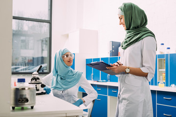 female muslim scientists using microscope and clipboard during experiment in chemical laboratory
