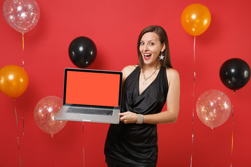 Excited young woman in black dress celebrating, holding laptop pc computer with blank black empty screen on bright red background air balloons. Happy New Year, birthday mockup holiday party concept.