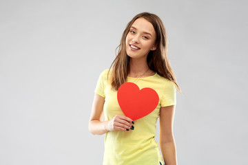 valentine's day and people concept - smiling young woman or teenage girl in yellow t-shirt with red heart over grey background