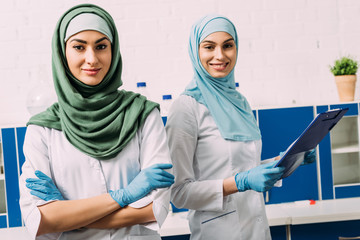beautiful female muslim chemists in hijab with arms crossed and clipboard looking at camera in laboratory