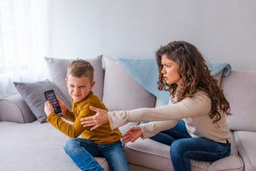 Boy having a tantrum and fighting with his mother for a smart phone sitting on a couch in the...