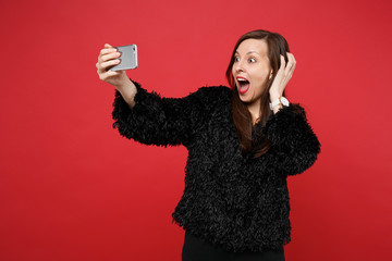 Excited young woman in black fur sweater clinging to head, doing selfie shot on mobile phone isolated on bright red wall background. People sincere emotions, lifestyle concept. Mock up copy space.