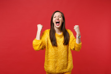 Screaming young girl in yellow fur sweater with closed eyes clenching fists like winner isolated on bright red wall background in studio. People sincere emotions lifestyle concept. Mock up copy space.