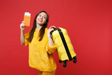 Pretty young woman in yellow fur sweater looking aside, holding suitcase, passport boarding pass ticket isolated on red wall background. People sincere emotions, lifestyle concept. Mock up copy space.