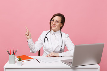 Beautiful female doctor sits at desk works on computer with medical document in hospital isolated on pastel pink wall background. Woman in medical gown glasses stethoscope. Healthcare medicine concept