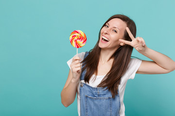 Portrait of cheerful young woman in denim clothes showing victory sign, holding colorful round lollipop isolated on blue turquoise background in studio. People lifestyle concept. Mock up copy space.