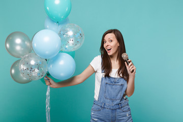 Fototapeta premium Excited young woman in denim clothes sing song in microphone, celebrating and holding colorful air balloons isolated on blue turquoise wall background. Birthday holiday party, people emotions concept.