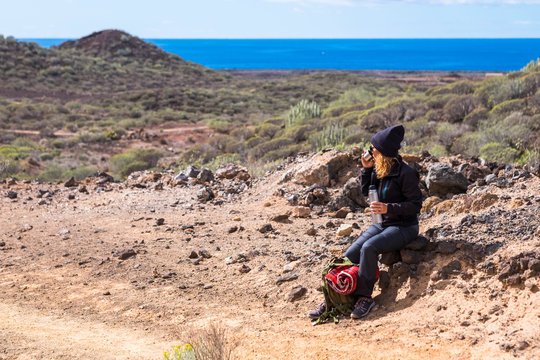 Curly cute woman doing trekking and hking in rock mountains with ocean in background - resting with drink - backpacker lifestyle for alternative leisure activity - people in outdoor