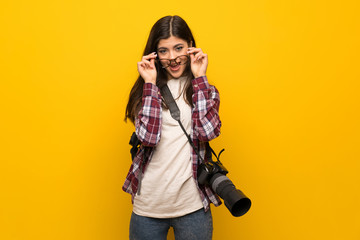 Photographer teenager girl over yellow wall with glasses and surprised