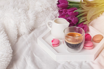 A cup of coffee, macarons and tulips on the white bedsheets.