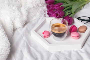 A cup of coffee, macarons and tulips on the white bedsheets.