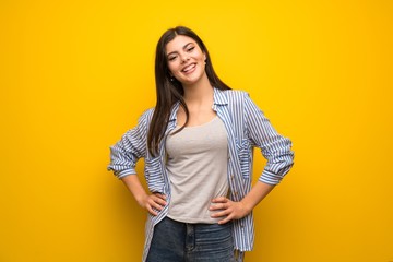 Teenager girl over yellow wall posing with arms at hip and smiling