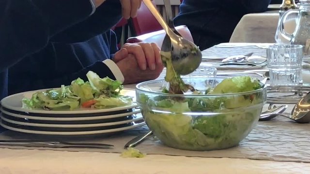 The Man Puts The Salad In A Plate. In The Dining Room, At The Table With Other People, Video Close-up