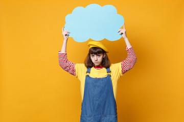 Upset puzzled girl teenager in french beret, denim sundress hold blue empty blank Say cloud, speech bubble isolated on yellow background. People sincere emotions lifestyle concept. Mock up copy space.