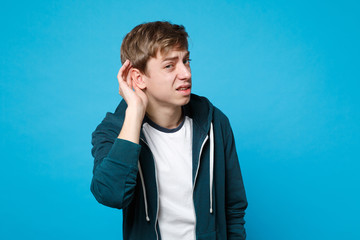 Portrait of curious young man in casual clothes eavesdrop with hearing gesture isolated on blue...