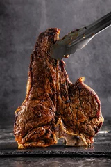 Gartenposter Large beef roast T-bone steak with smoke. Head chef holding steak meat tongs on a black background. photon image. copy space © zukamilov