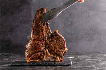 Large beef roast T-bone steak with smoke. Head chef holding steak meat tongs on a black background....