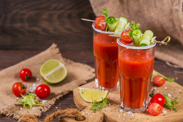Two glasses of fresh organic tomato juice decorated with raw tomatoes, cucumber and leaves on a rustic wooden cutting board