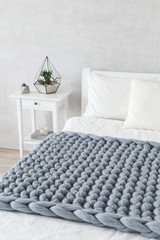 Grey knitted woolen merino chunky blanket. Thick yarn. Stylish cozy scandinavian bedroom interior: bed with white linen, bedside table, grey stucco or cement or plaster wall.